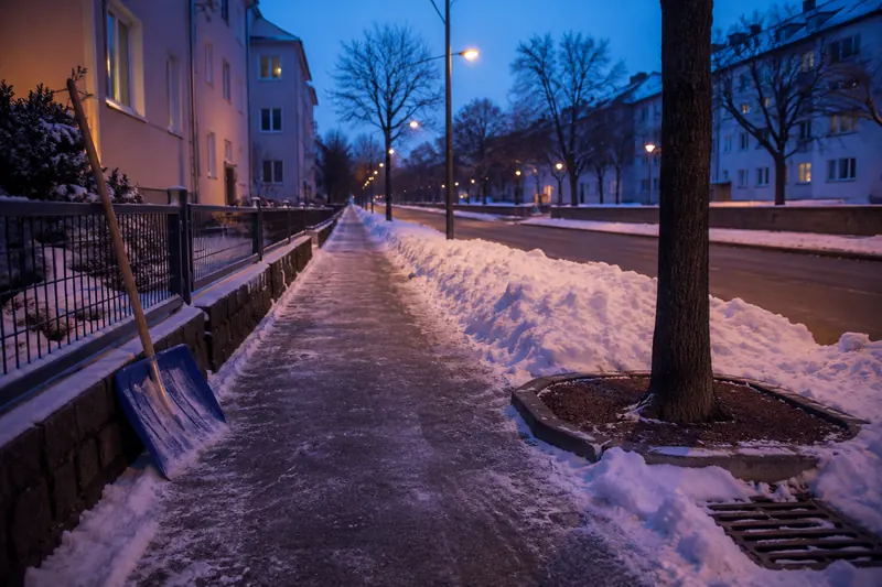 Geräumter Gehweg in der Dämmerung, der Schnee liegt am Fahrbahnrand aufgehäuft, Baumscheibe und Kanaleinlauf sind frei